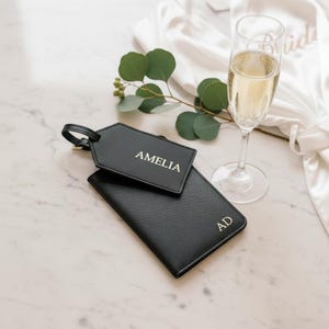 Könnte beinhalten: Ein schwarzer Kofferanhänger mit dem in Gold geprägten Namen AMELIA und ein passender schwarzer Reisepasshalter mit den goldenen Initialen AD. Die Gegenstände sind neben einem Glas Champagner und einem Zweig Eukalyptusblättern abgebildet.