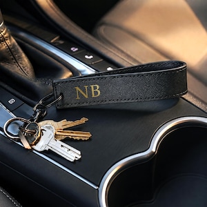 Könnte beinhalten: Ein schwarzes Schlüsselanhängerband aus strukturiertem Leder mit den gold geprägten Buchstaben NB. Das Band ist an einem schwarzen Metallring und einem schwarzen Metallkarabiner befestigt, an dem drei silber- und messingfarbene Schlüssel hängen.
