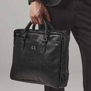 May include: Black leather briefcase with a top handle and a zippered closure. The briefcase has the initials "CL" on the front. The bag is held by a person wearing a gray blazer and black pants.