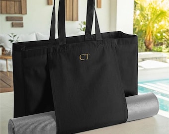 May include: A black canvas tote bag featuring two long shoulder straps and the gold embroidered letters CT centred on the front. A rolled-up light grey yoga mat is inserted horizontally through the bottom section of the bag.