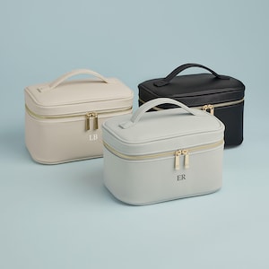 May include: Three rectangular cosmetic cases with top handles and gold-toned zippers. One case is beige with the letters LB, one is light gray with the letters ER, and one is black. Each case features a smooth finish and a structured design.