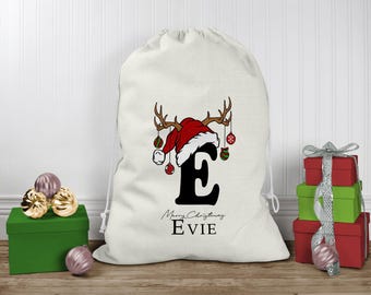 May include: A white fabric drawstring gift sack featuring a large black letter E topped with a red and white Santa hat and brown reindeer antlers. Hanging from the antlers are small colorful ornaments. The text reads Merry Christmas EVIE.
