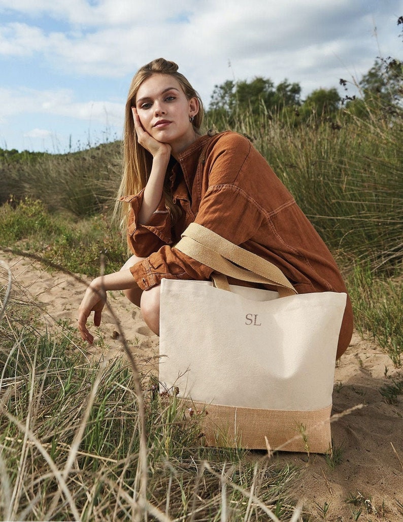 Pu&ograve; includere: Una borsa tote in tela bianca con fondo in juta marrone. La borsa ha una tracolla marrone e le iniziali "SL" ricamate sul davanti.