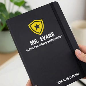 Può includere: Un taccuino nero con chiusura elastica caratterizzato da un'icona a scudo gialla con una stella nera. Il testo MR. EVANS PLANS FOR WORLD DOMINATION* e *AND ALSO LESSONS è stampato in lettere maiuscole bianche sulla copertina del taccuino.