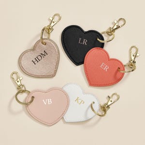 May include: Five heart-shaped keychains in various colors: gold, black, coral, pink, and white. Each keychain has a gold-colored clasp and ring. The keychains are personalized with initials: HDM, LR, ER, VB, and KP.