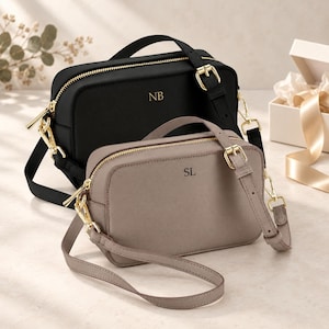 May include: Two rectangular crossbody bags with gold-tone hardware and adjustable straps. The black bag features the initials NB, while the taupe bag features the initials SL. Both bags have top zippers and a structured design with matching shoulder straps.
