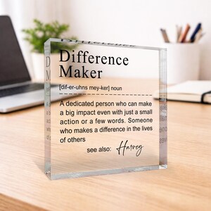 A clear glass block with a definition of a " Difference Maker" written on it, placed on a wooden desk with a laptop and a plant in the background.