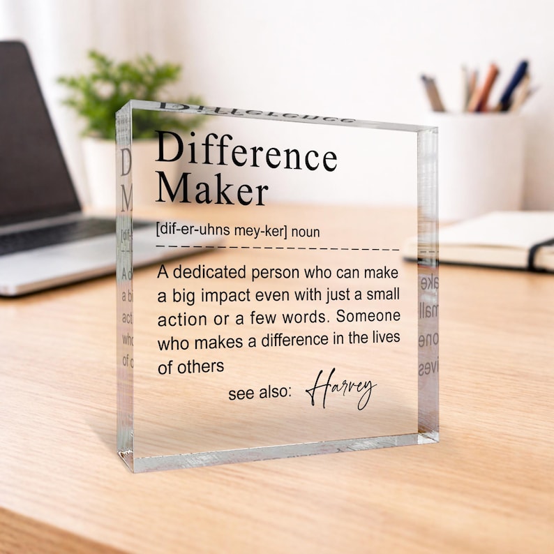 A clear glass block with a definition of a " Difference Maker" written on it, placed on a wooden desk with a laptop and a plant in the background.