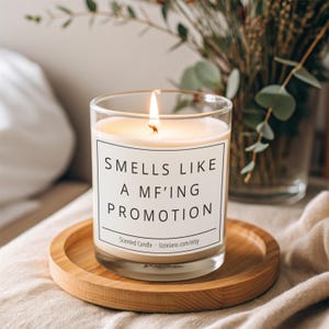 Peut inclure: Une bougie en pot de verre transparent avec une étiquette blanche portant le texte noir 'SMELLS LIKE A MF'ING PROMOTION' et 'Scented Candle lizzielane.com/etsy'. La bougie a une mèche allumée et repose sur un sous-verre en bois rond.