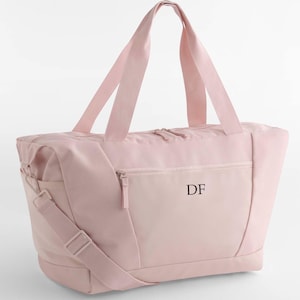 Peut inclure: Un sac de sport rose clair avec une bandoulière et deux poignées. Le sac a une poche avant zippée et les initiales "DF" imprimées en noir. Le sac est fait d'un matériau doux et durable.