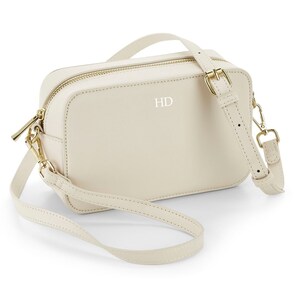 May include: A cream-colored leather crossbody bag with a gold zipper and a gold buckle. The bag has a short strap and a long adjustable strap. The initials "HD" are embossed on the front of the bag.