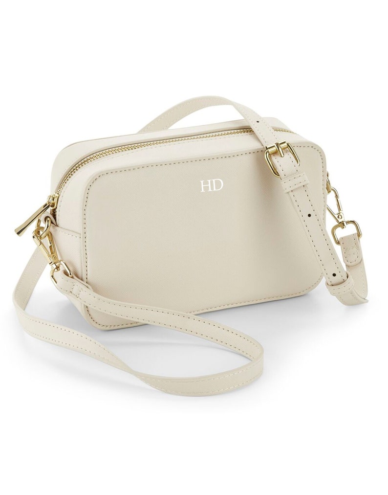 May include: A cream-colored leather crossbody bag with a gold zipper and a gold buckle. The bag has a short strap and a long adjustable strap. The initials "HD" are embossed on the front of the bag.