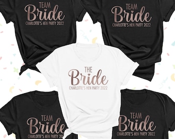 May include: Four black t-shirts and one white t-shirt with short sleeves and crew necks. The black shirts feature the rose gold text TEAM Bride and CHARLOTTE'S HEN PARTY 2022. The white shirt features the rose gold text THE Bride and CHARLOTTE'S HEN PARTY 2022.