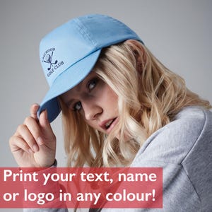 May include: A light blue baseball cap featuring a dark blue embroidered logo on the front. The logo depicts a golf ball and two crossed golf clubs with the text LAKEWOOD GOLF CLUB arched above and below the graphic in a circular arrangement.