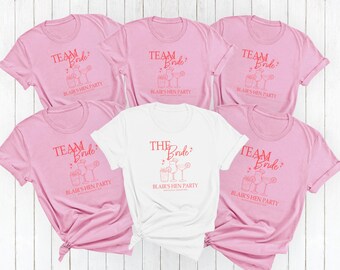 May include: Five short-sleeved crew neck t-shirts arranged in a group. Four shirts are light pink with red text reading TEAM BRIDE and BLAIR'S HEN PARTY above cocktail illustrations. One central white shirt reads THE BRIDE and BLAIR'S HEN PARTY.