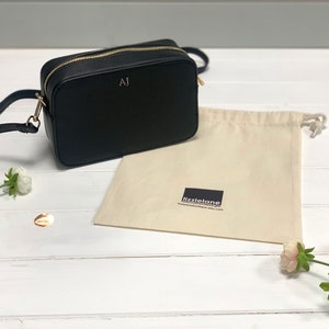May include: A black leather crossbody bag with a gold zipper and the initials "AJ" embossed on the front. The bag is sitting on a white wooden surface with a white cotton drawstring bag and flowers.