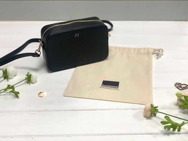 May include: A black leather crossbody bag with a gold zipper and the initials "AJ" embossed on the front. The bag is sitting on a white wooden surface with a white cotton drawstring bag and flowers.