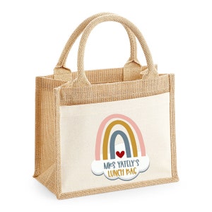 May include: A beige burlap lunch bag with a white pocket featuring a rainbow design and the text "Mrs. Yately's Lunch Bag." The bag has two beige handles and is designed for carrying lunches. The rainbow includes pink, gold, and blue arches.