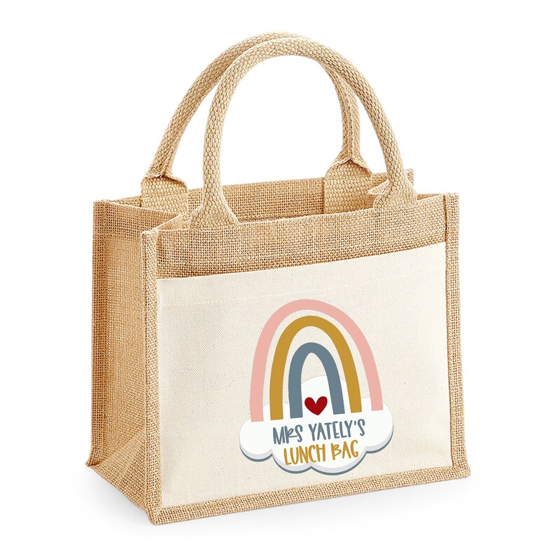 May include: A beige burlap lunch bag with a white pocket featuring a rainbow design and the text "Mrs. Yately's Lunch Bag." The bag has two beige handles and is designed for carrying lunches. The rainbow includes pink, gold, and blue arches.