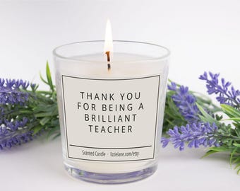 Op de afbeelding: Een brandende geurkaars in een heldere glazen pot met een wit etiket met de zwarte tekst THANK YOU FOR BEING A BRILLIANT TEACHER en lizzielane.com/etsy, geflankeerd door takjes paarse lavendelbloemen op een effen witte achtergrond.