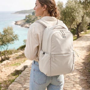 May include: A beige backpack featuring a top handle, adjustable shoulder straps, and a front zippered pocket. The upper portion of the bag displays the embroidered letters RE in a dark font. The design includes side pockets with drawstring closures.