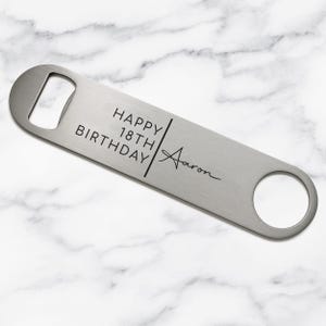 May include: A flat, rectangular stainless steel bottle opener with a brushed metal finish. The surface is engraved with the text HAPPY 18TH BIRTHDAY separated by a vertical line from the name Aaron written in a cursive script font.