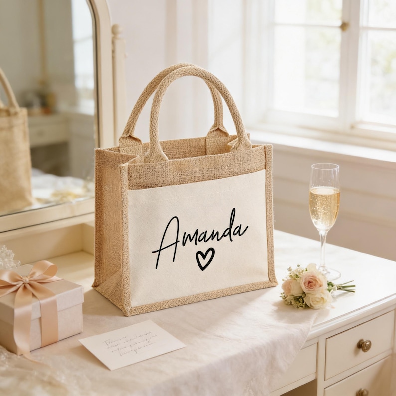 a beige tote bag with the name "Amanda" printed on it, placed on a white dresser next to a bouquet of flowers and a glass of champagne.