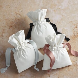 May include: Three white fabric drawstring bags with different colored ribbons: black, light gray, and dusty rose. The bags are gathered at the top and tied with the ribbons. The background is a mottled beige.