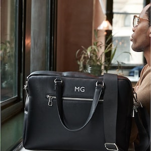 May include: Black leather briefcase with silver zippers, handles, and a shoulder strap. The initials "MG" are displayed on the front. The bag is sitting on a white surface near a window.