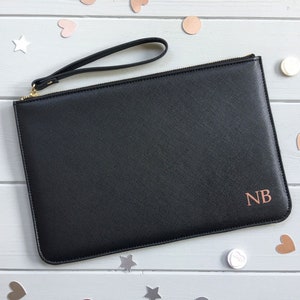 May include: Black leather clutch with a gold zipper and a black wrist strap. The clutch has a rose gold monogram "NB" on the front.