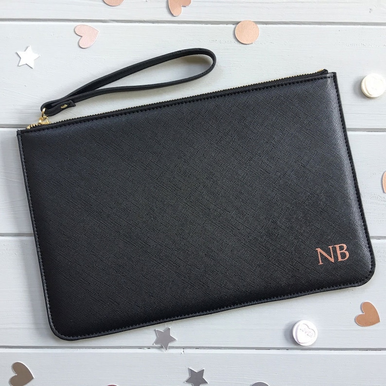 May include: Black leather clutch with a gold zipper and a black wrist strap. The clutch has a rose gold monogram "NB" on the front.