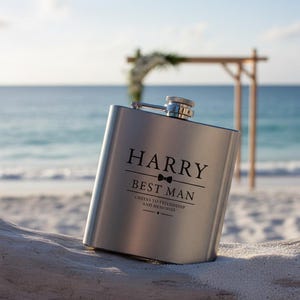 Może przedstawiać: Srebrny flakon z grawerunkiem "HARRY BEST MAN". Flakon spoczywa na piaszczystej powierzchni, w tle morze i drewniany łuk. Flakon ma srebrną nakrętkę. Wygrawerowano również tekst "Cheers to friendship and memories".