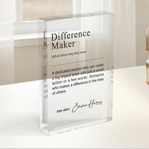 a clear glass display case with a definition of a " Difference Maker" written on it. The display case is placed on a table, and there is a vase of flowers nearby.