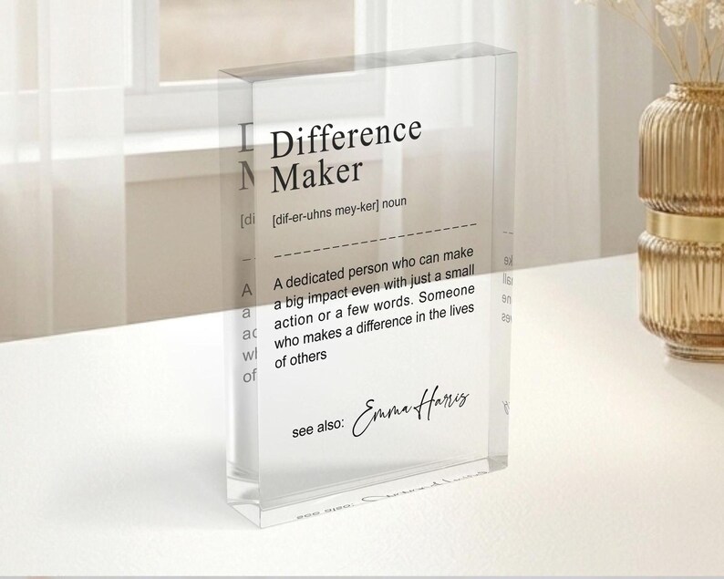 a clear glass display case with a definition of a " Difference Maker" written on it. The display case is placed on a table, and there is a vase of flowers nearby.