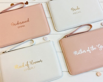 May include: Four pink and white clutch purses with gold and white lettering. The purses say "Bride Abigail", "Bridesmaid Emma", "Maid of Honour Kimberly", and "Mother of the Bride".