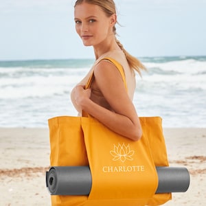 A woman stands on a beach, holding a large yellow tote bag with a yoga mat inside.