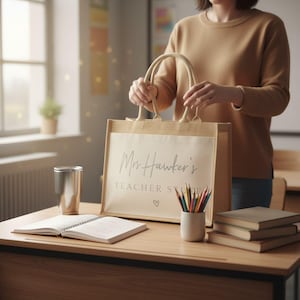 A woman holding a bag with the text "Mrs Hawker's Teacher Stuff" on it, standing in front of a desk with books, a cup, and a pencil holder.