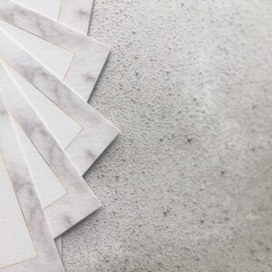 May include: A stack of white paper cards with a gold border and a marble pattern. The cards are arranged in a fan shape on a gray textured surface.