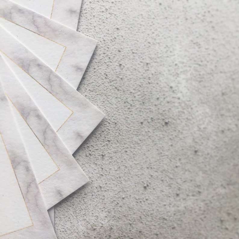 May include: A stack of white paper cards with a gold border and a marble pattern. The cards are arranged in a fan shape on a gray textured surface.