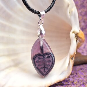 May include: A pendant necklace with a purple resin teardrop-shaped pendant. The pendant has a black heart design and is suspended from a silver bail and a black cord. The pendant is approximately 1.5 inches long.