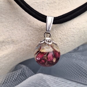 May include: A black choker necklace with a silver pendant. The pendant is a clear orb filled with dried red and white flowers. The orb is attached to a silver bail and a black suede cord.