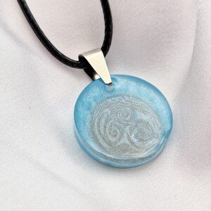 May include: A round pendant with a light blue resin finish and a silver-colored Celtic knot design. The pendant is attached to a black cord necklace with a silver-colored clasp.