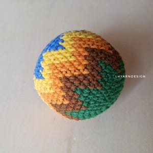 Puede incluir: Una pelota esférica tejida a mano con un vibrante patrón de chevrón multicolor en tonos azul, amarillo, naranja, marrón y verde. El texto LHYARNDESIGN está impreso en blanco en el lado derecho de este objeto texturizado hecho de hilo.
