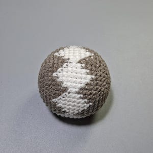Puede incluir: Una pelota de ganchillo esférica con una superficie texturizada hecha de hilo marrón. La pelota está decorada con un patrón de punto de cruz blanco que forma una figura vertical en forma de diamante que recorre el centro del objeto.