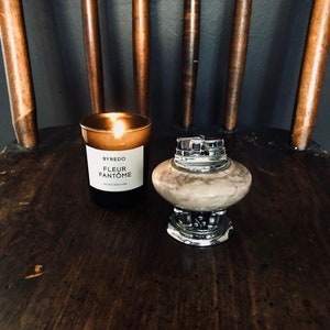 May include: A brown glass candle with a white label that reads "Byredo Fleur Fantome" on a wooden surface. Next to the candle is a silver and marble cigarette lighter.