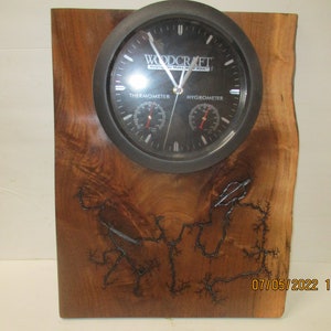 Könnte beinhalten: Eine schwarze Uhr mit Thermometer und Hygrometer auf einem Holzsockel mit einem Holzbrandmuster. Das Zifferblatt der Uhr trägt den Text "Woodcraft Helping You Make It Shine".