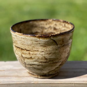 May include: A handmade ceramic bowl with a textured, light brown exterior and a darker brown rim. The bowl has a rounded shape and sits on a wooden surface, with a blurred green background.