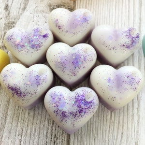 May include: Seven white heart-shaped wax melts with purple glitter embedded in the wax. The melts are arranged in a circle on a light wood surface.