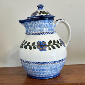 Puede incluir: Una jarra de cerámica con tapa, que presenta una base blanca decorada con patrones florales azules y verdes, una banda central de flores y hojas, y bordes pintados con esponja azul alrededor del cuello, el asa y la base del recipiente.