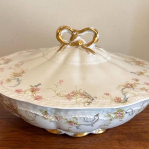 May include: A white porcelain serving dish with a lid, featuring a floral pattern in pink and green. The dish has gold accents on the handles and a bow-shaped handle on the lid. The dish is on a wooden surface.